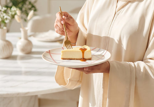A person in a cream-colored robe holds a white plate with a slice of cheesecake topped with caramel sauce, using a gold fork, in a bright, modern kitchen setting.